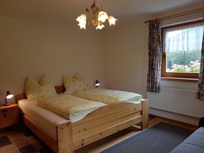 A bedroom with a wooden bed, patterned pillows, a nightstand with a lamp, a window with curtains, and a chandelier. - Ferienhaus-Sonnblick (Sillian)