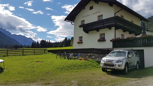 Un edificio de dos pisos con un balcón, un coche estacionado y montañas al fondo.