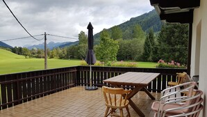 Un patio con una mesa y sillas de madera, un paraguas negro, y vistas a un campo verde y montañas.