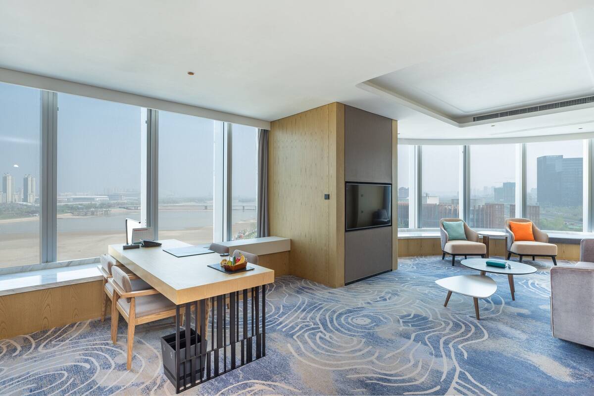 Ein modernes Hotelzimmer mit großem Fenster, einem Holztisch, einem Flachbildfernseher und einer Sitzecke mit zwei Stühlen und einem kleinen Tisch.