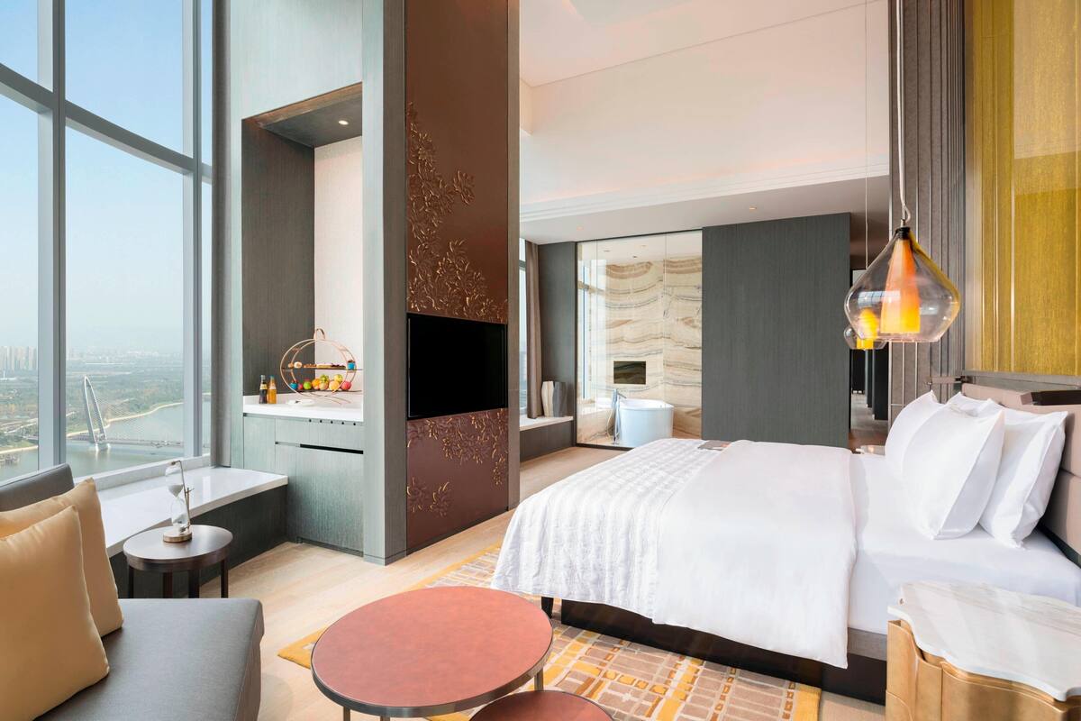 Ein modernes Hotelzimmer mit einem großen Bett, einem an der Wand befestigten Fernseher, einem kleinen Tisch und Blick auf die Stadt durch große Fenster.