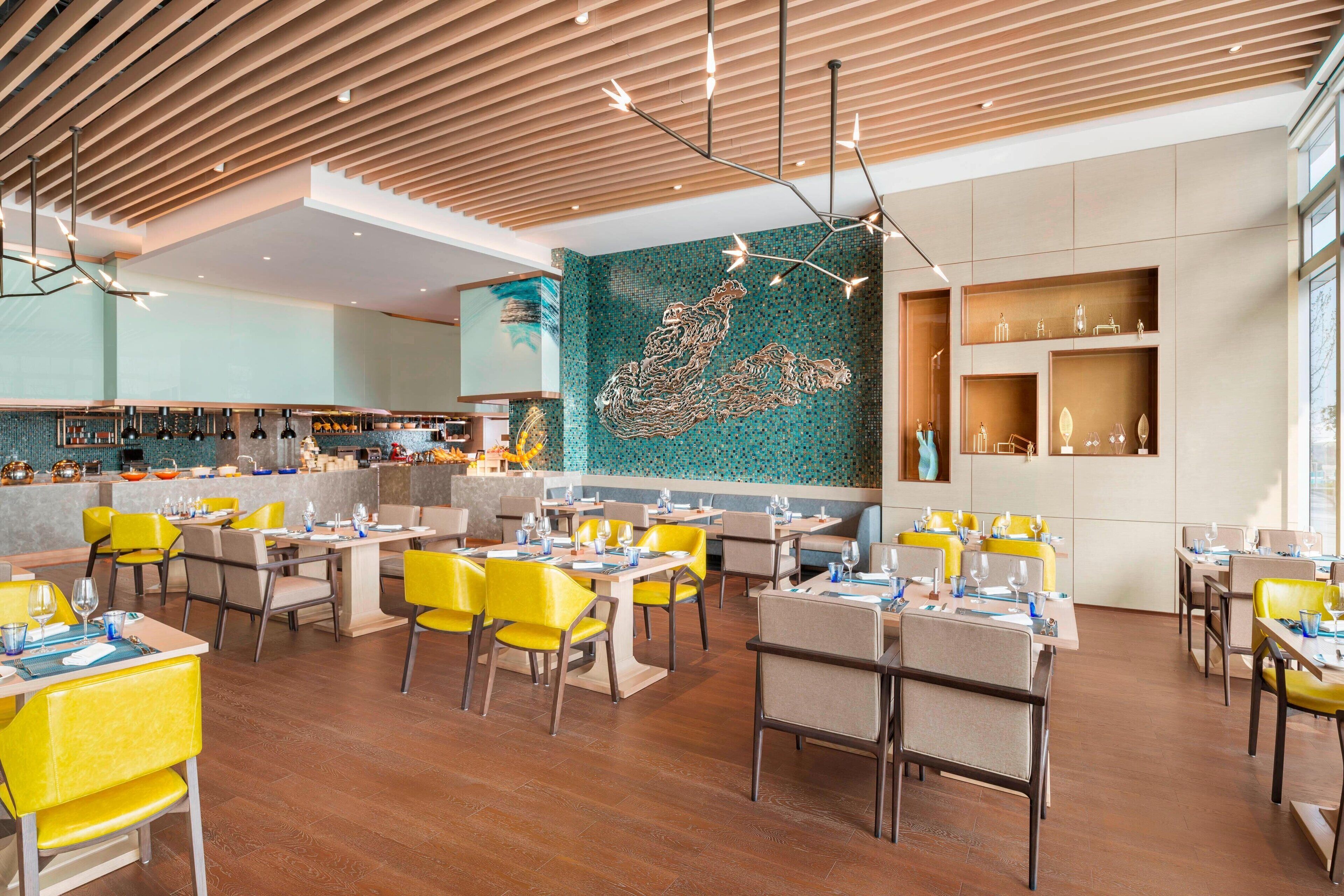 Interior de un restaurante moderno con sillas amarillas, techo de madera y una obra de arte mural de mosaico.