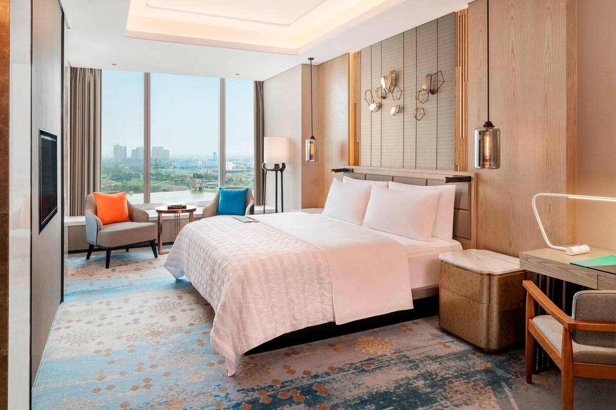 Ein Hotelzimmer mit einem großen Bett, einem Schreibtisch, einem Sessel, einem Fernseher und Blick auf die Stadt.