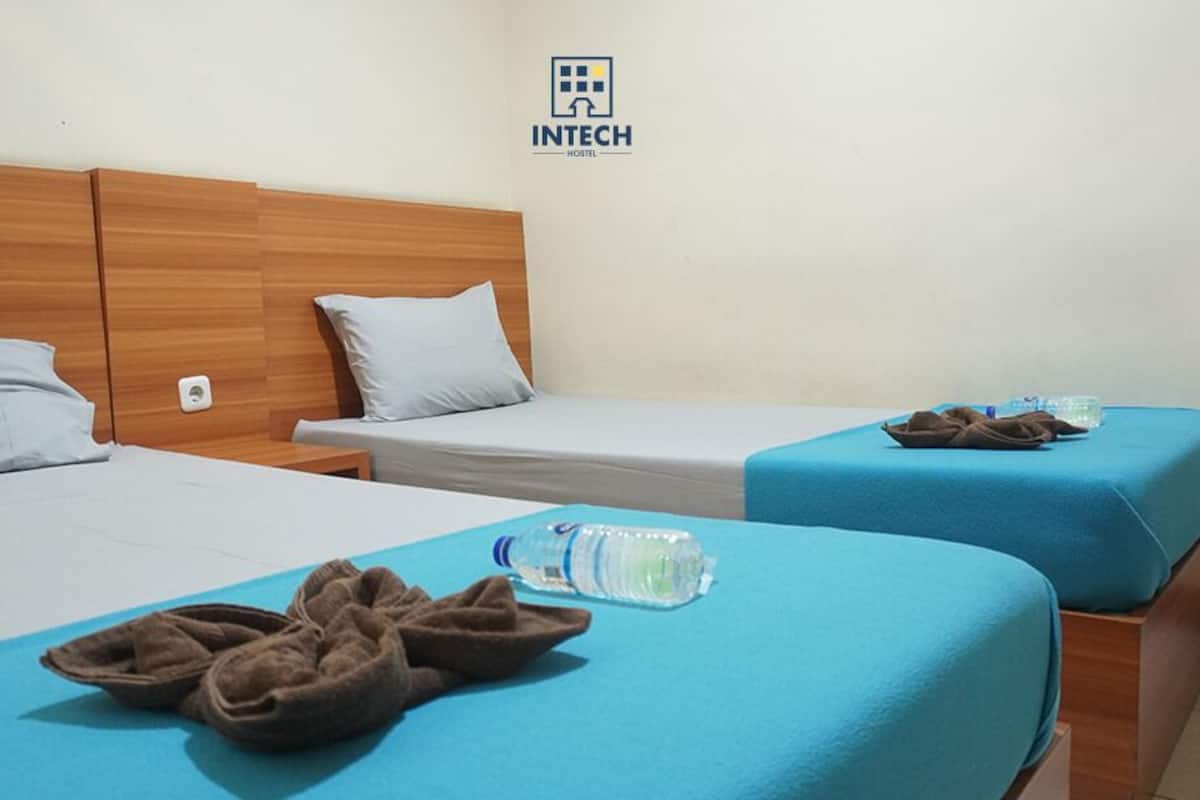 A hotel room with two beds, each with a blue bedspread and pillows. There is a water bottle and slippers on the beds.