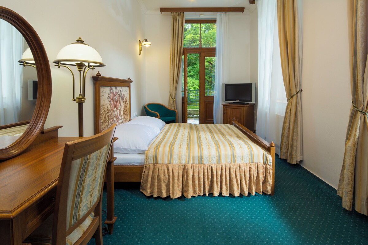 A hotel room with a bed, a desk with a mirror, a chair, a television, and a window with curtains.