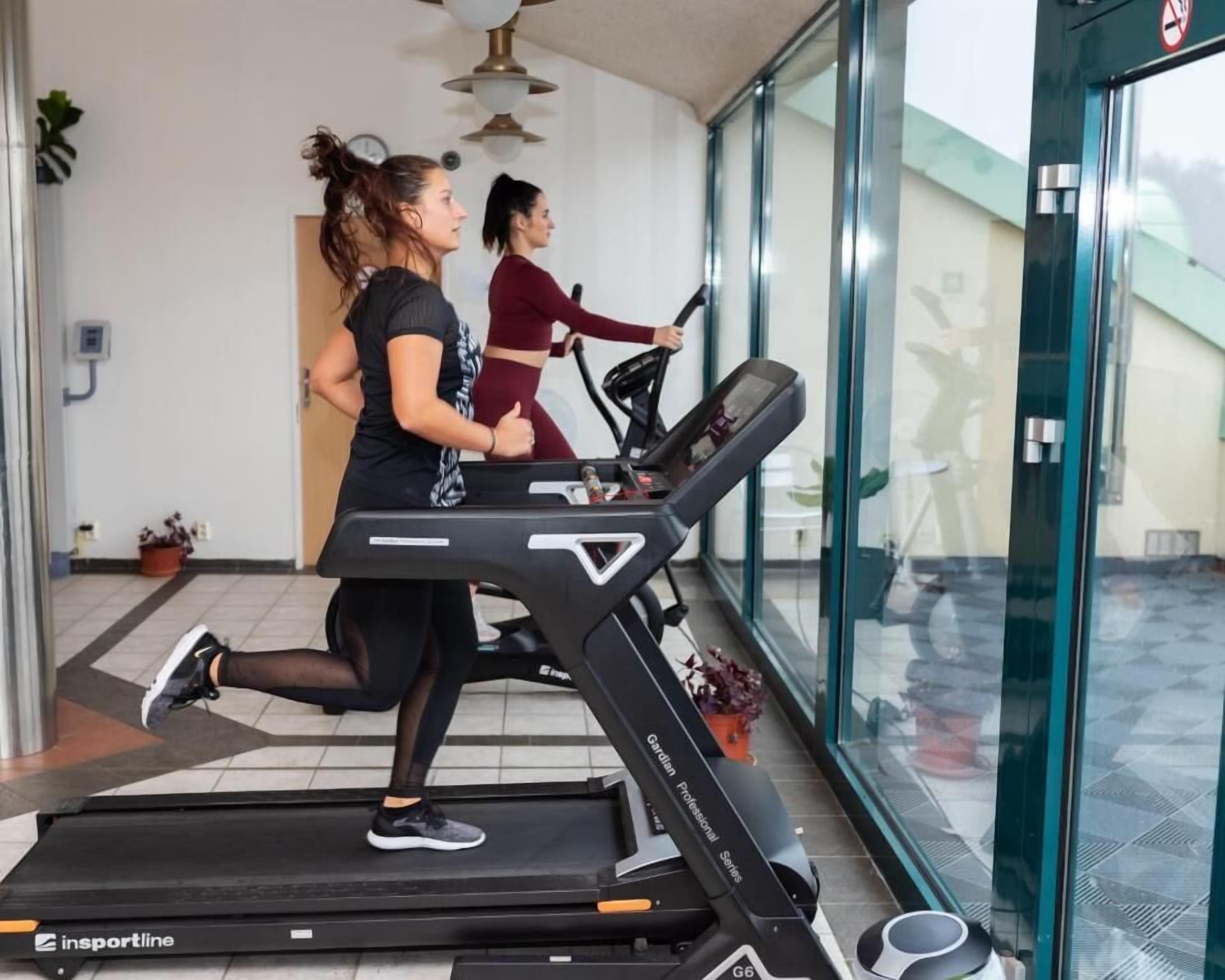 Two individuals running on a treadmill in a modern indoor setting with glass walls and a ceiling fan.
