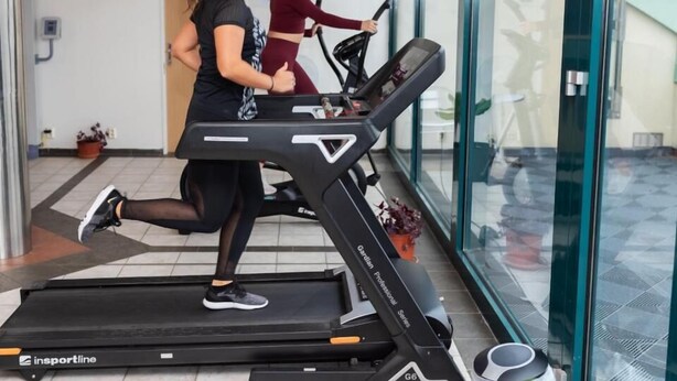 Dos personas corriendo en una cinta de correr en un espacio interior moderno con paredes de vidrio y un ventilador de techo.