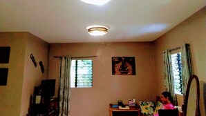 A room with two ceiling lights, a window with blinds, a small table with items, and a chair.