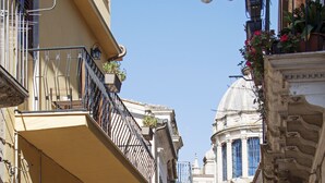A narrow street with buildings featuring balconies, a dome structure in the background, and a clear blue sky. - BFAME - Cornice Barocca (Ragusa)
