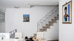 A dining area with a table set for two, a coffee pot, pastries, and a potted plant. A staircase with a railing is visible in the background. - BFAME - Cornice Barocca (Ragusa)