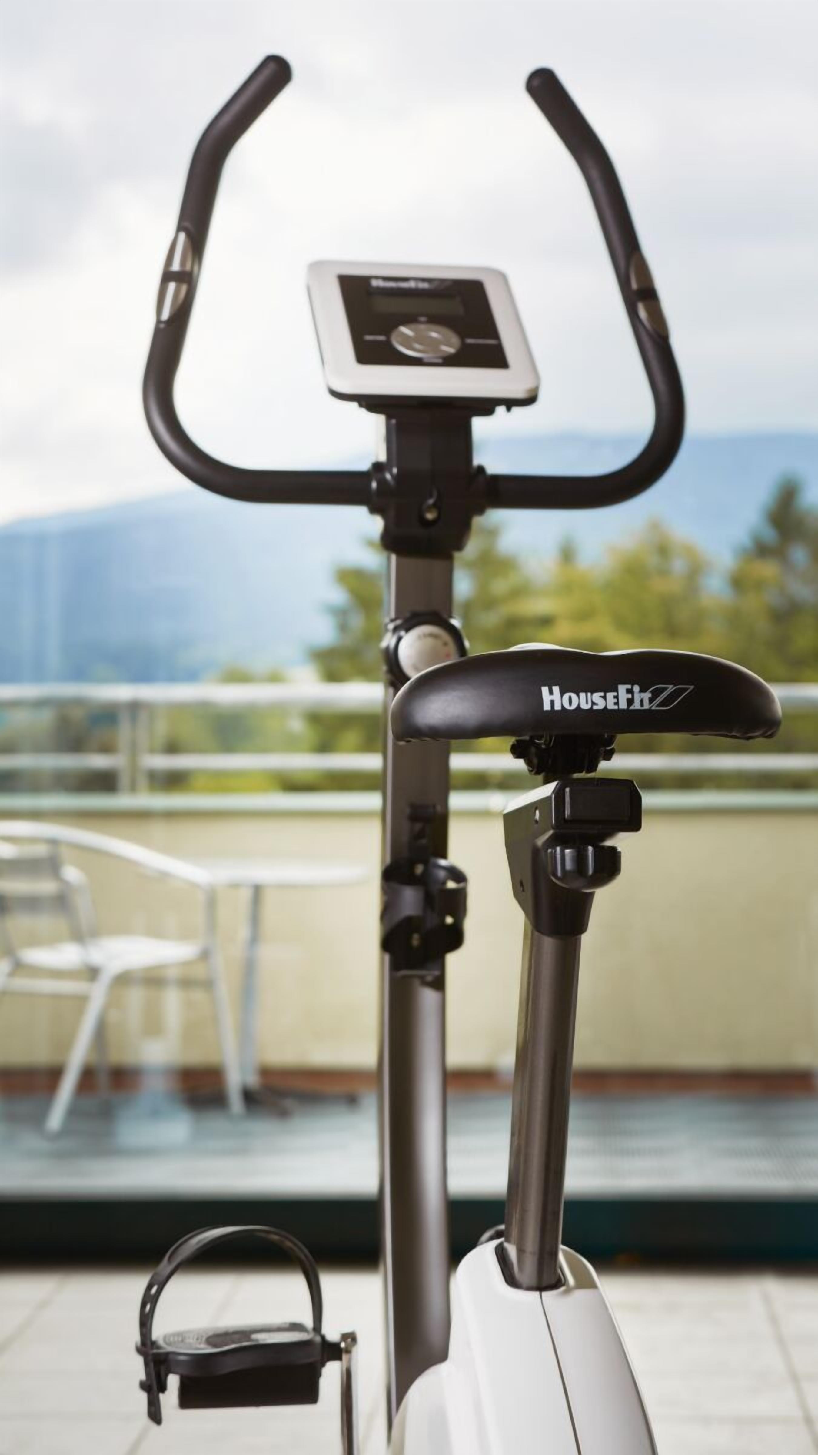 En motionscykel med en digital display och ett säte med ett varumärkeslogotyp, placerad utomhus med utsikt över ett räcke och grönska.