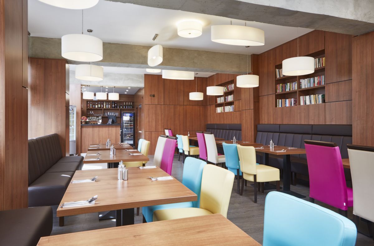 Een modern restaurant met houten lambrisering, kleurrijke stoelen en hanglampen.