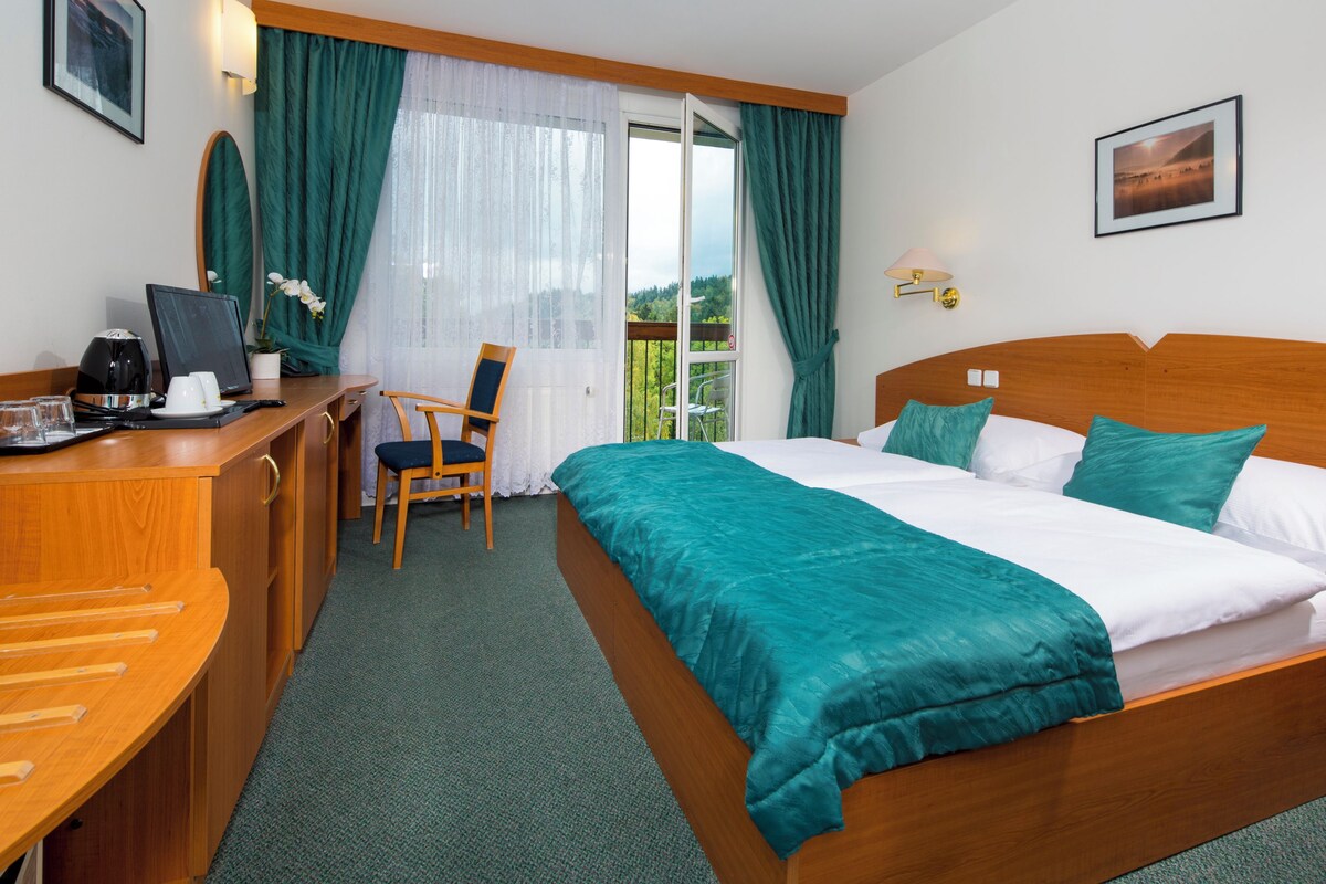 A hotel room with a bed, a desk with a computer, a chair, and a view of greenery through the window.