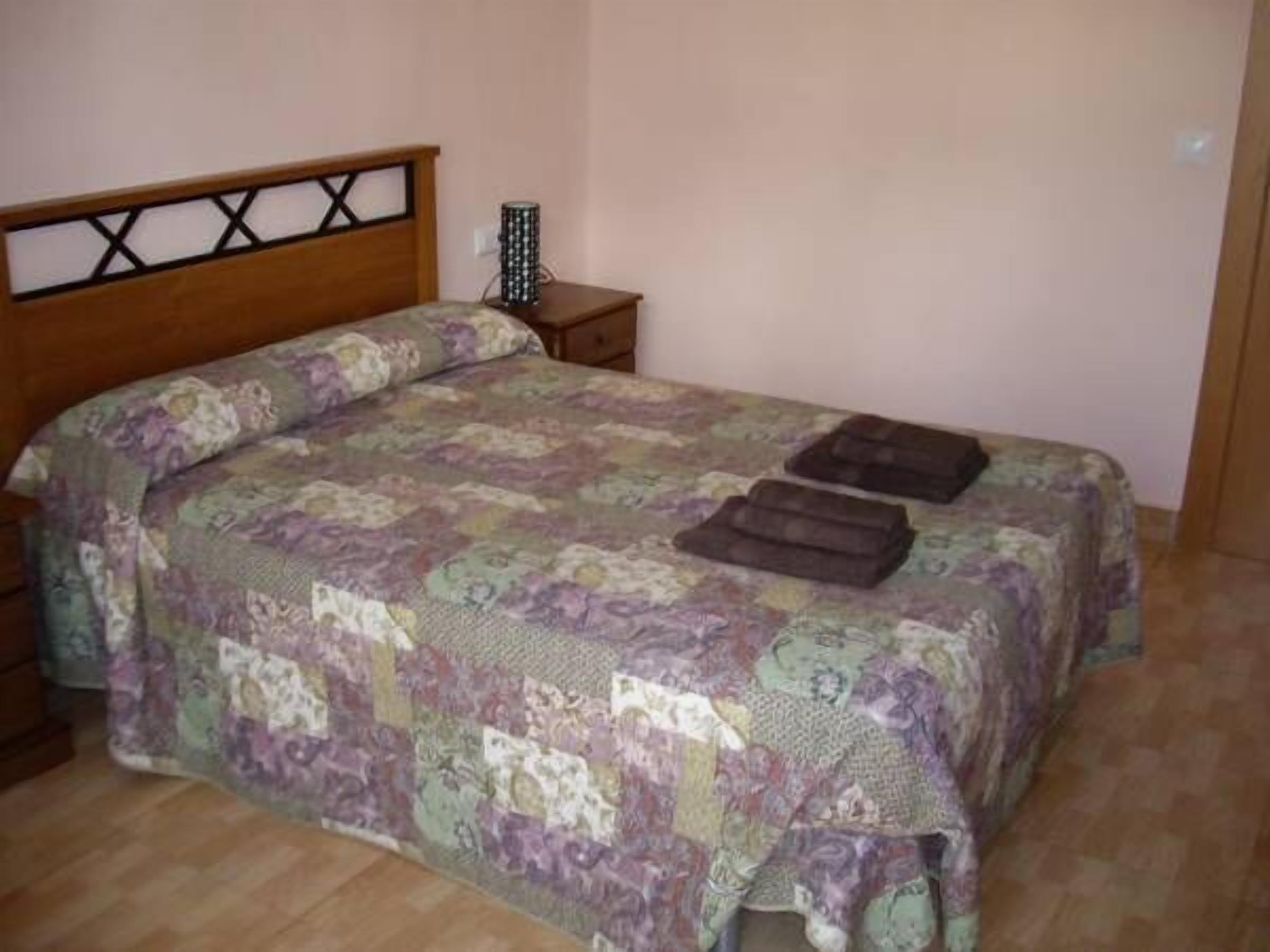 A bedroom with a wooden headboard, a patterned bedspread, two pillows, and a bedside table with a lamp.