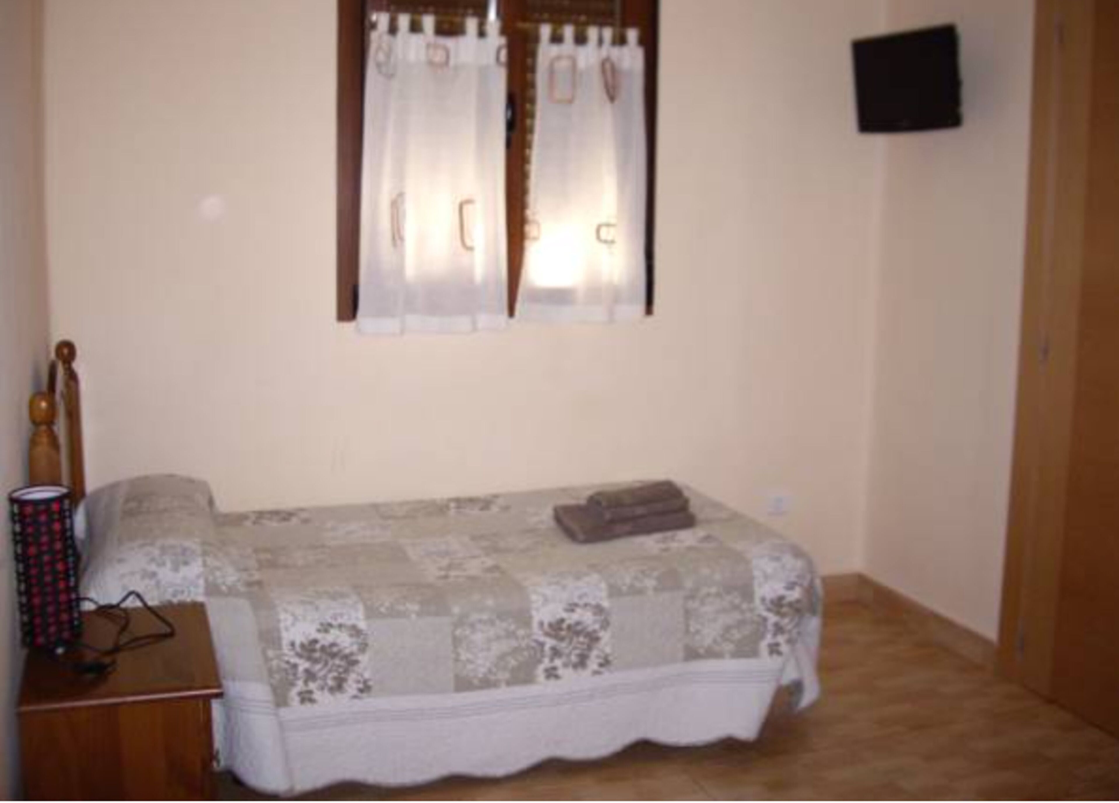 Un dormitorio pequeño con una cama, una mesita de noche de madera, una ventana con cortinas y un televisor montado en la pared.