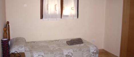 Un dormitorio pequeño con una cama, una mesita de noche de madera, una ventana con cortinas y un televisor montado en la pared.