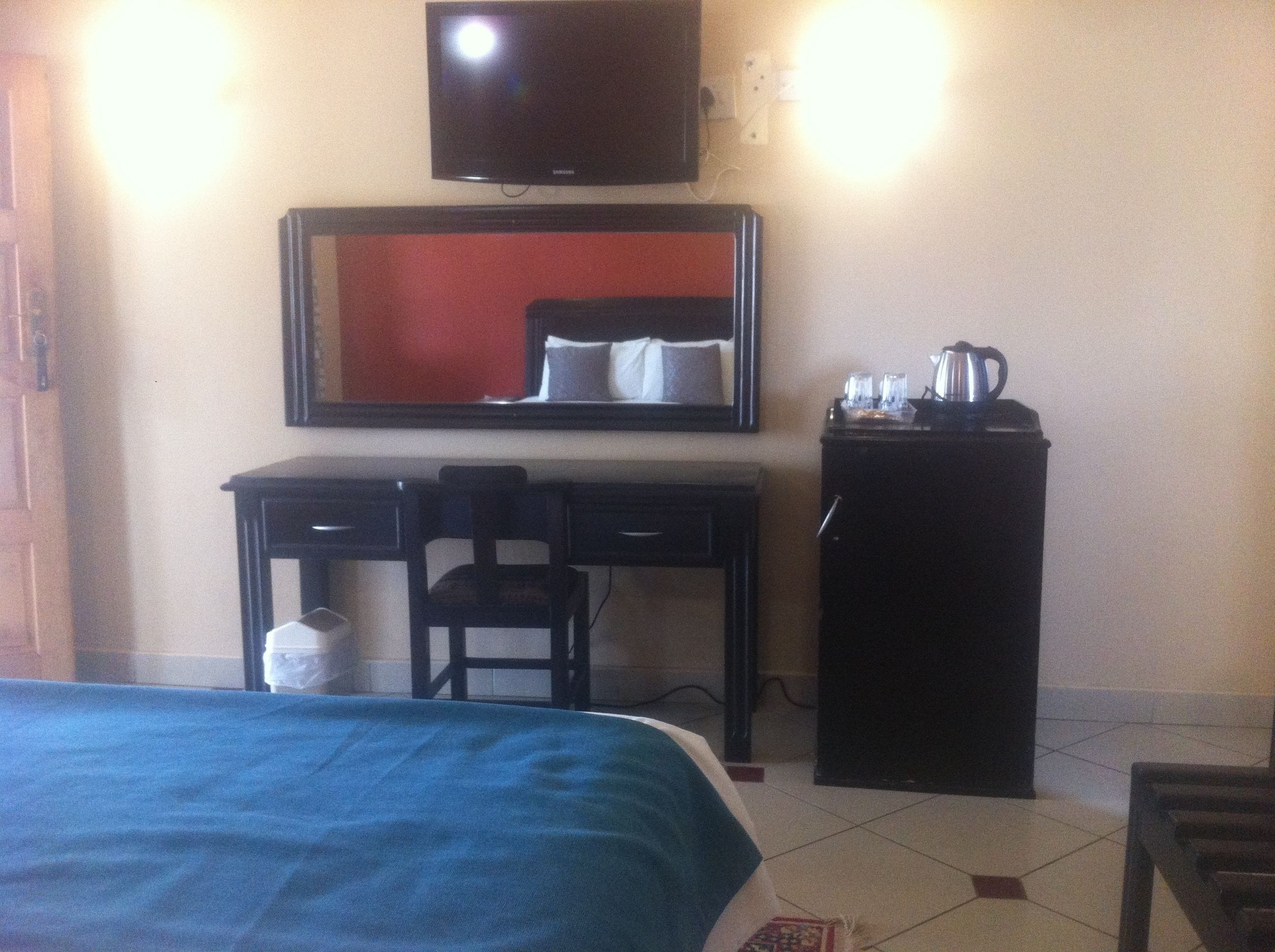 A hotel room with a bed, a desk with a chair, a television mounted on the wall, and a small cabinet with a kettle.