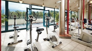 Une salle de sport équipée de vélos d’appartement et d’appareils de cardio, de grandes fenêtres offrant une vue sur les arbres et les montagnes, et d’un pilier rouge.