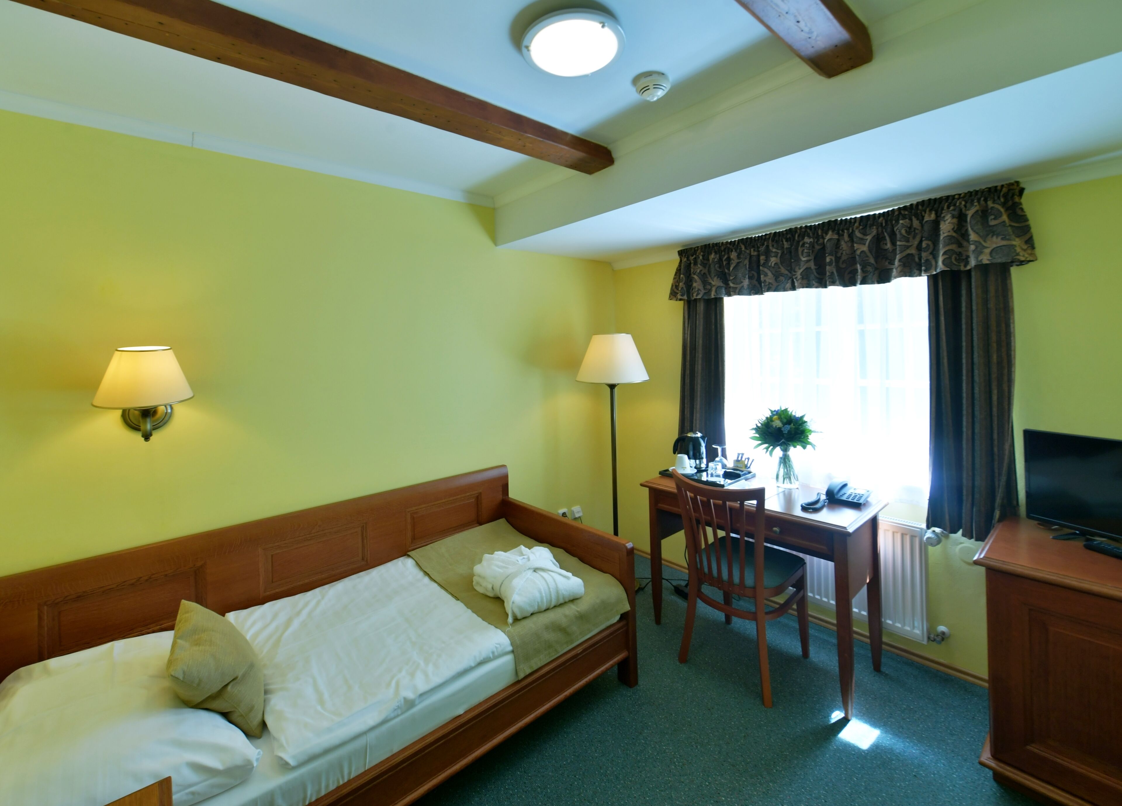 A hotel room with a bed, desk, chair, lamp, TV, and window with curtains.