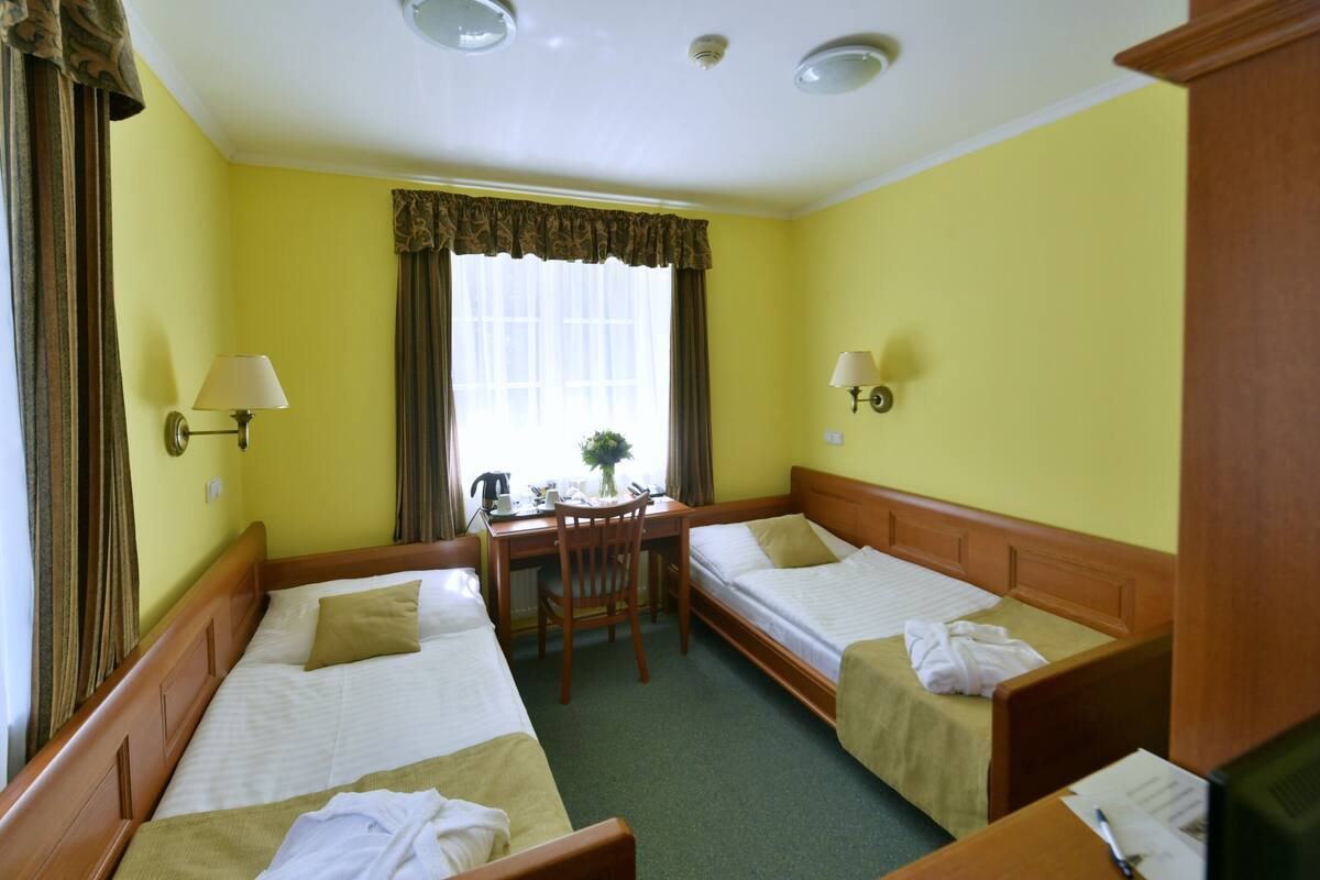 A hotel room with two single beds, a wooden desk, a chair, and a small table with a lamp.