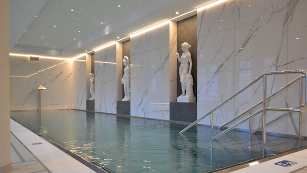 Une piscine intérieure aux murs de marbre et ornée de statues, avec une eau cristalline et un escalier en métal.