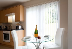 A modern kitchen with wooden cabinets, a glass table, white chairs, and a bottle of champagne on the table. - Beech Lodge (Oxford)