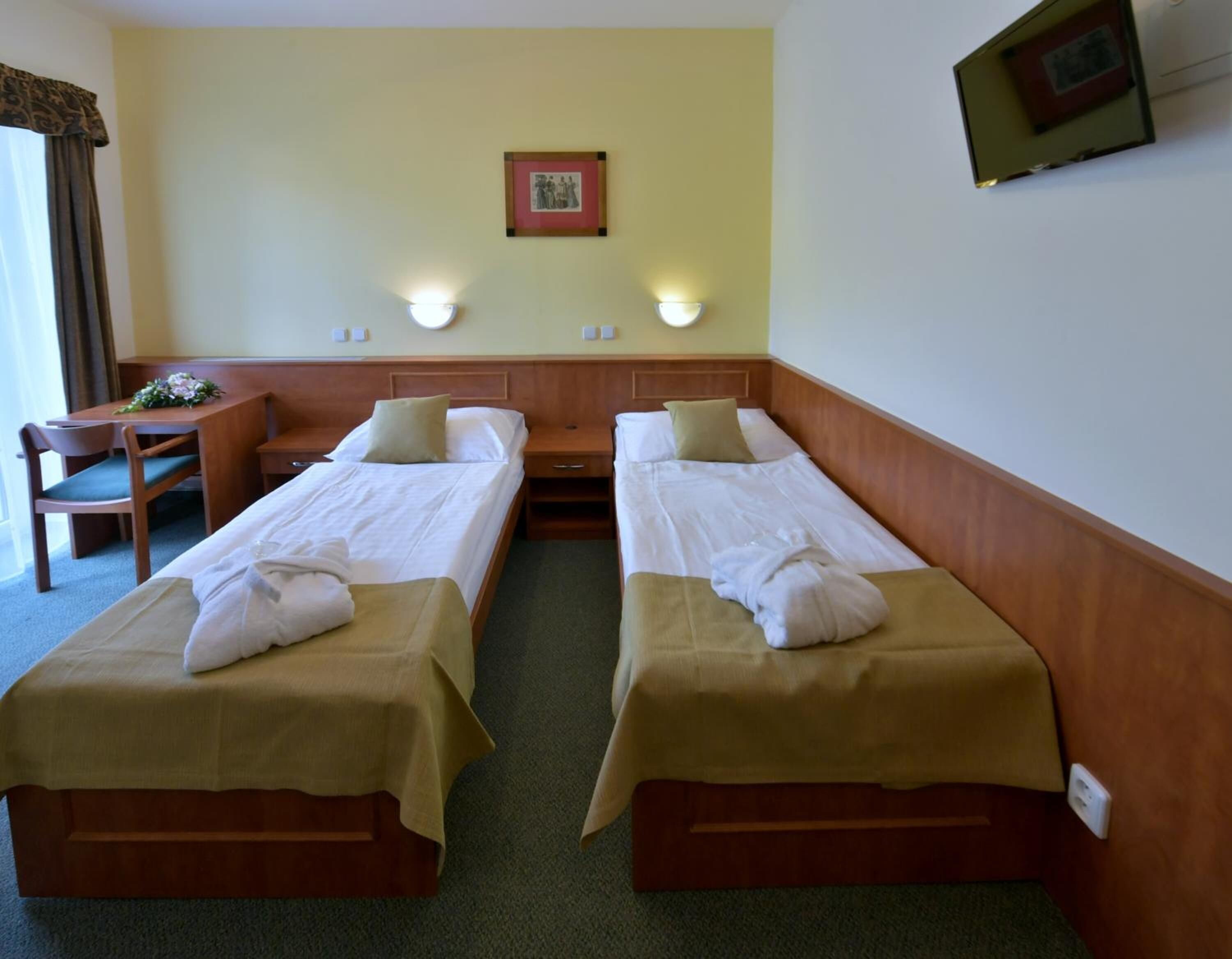 A hotel room with two single beds, a desk with a chair, and a small table with a vase of flowers.
