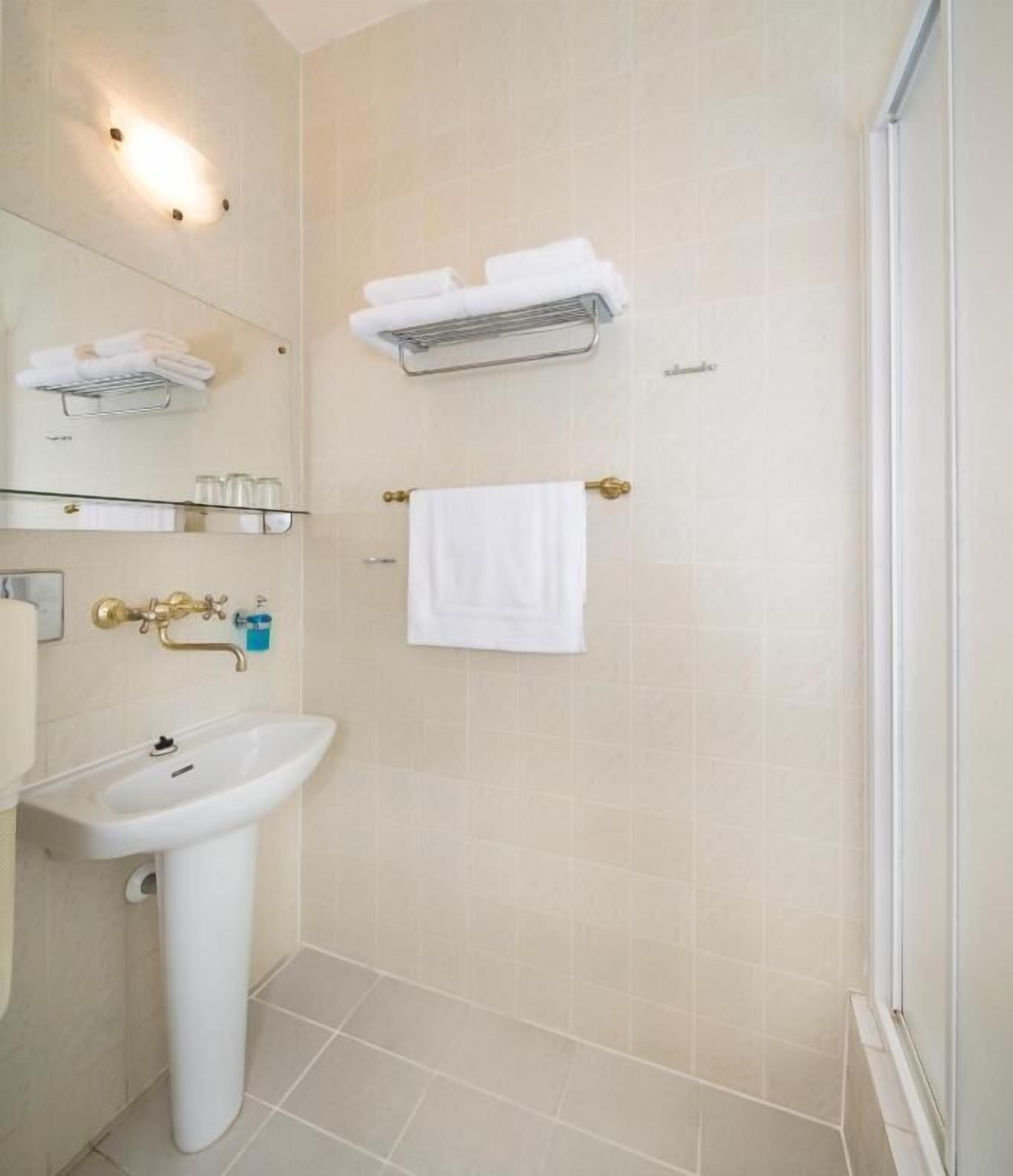 A bathroom with a pedestal sink, a towel rack with folded towels, a glass shelf with toiletries, and a shower area.