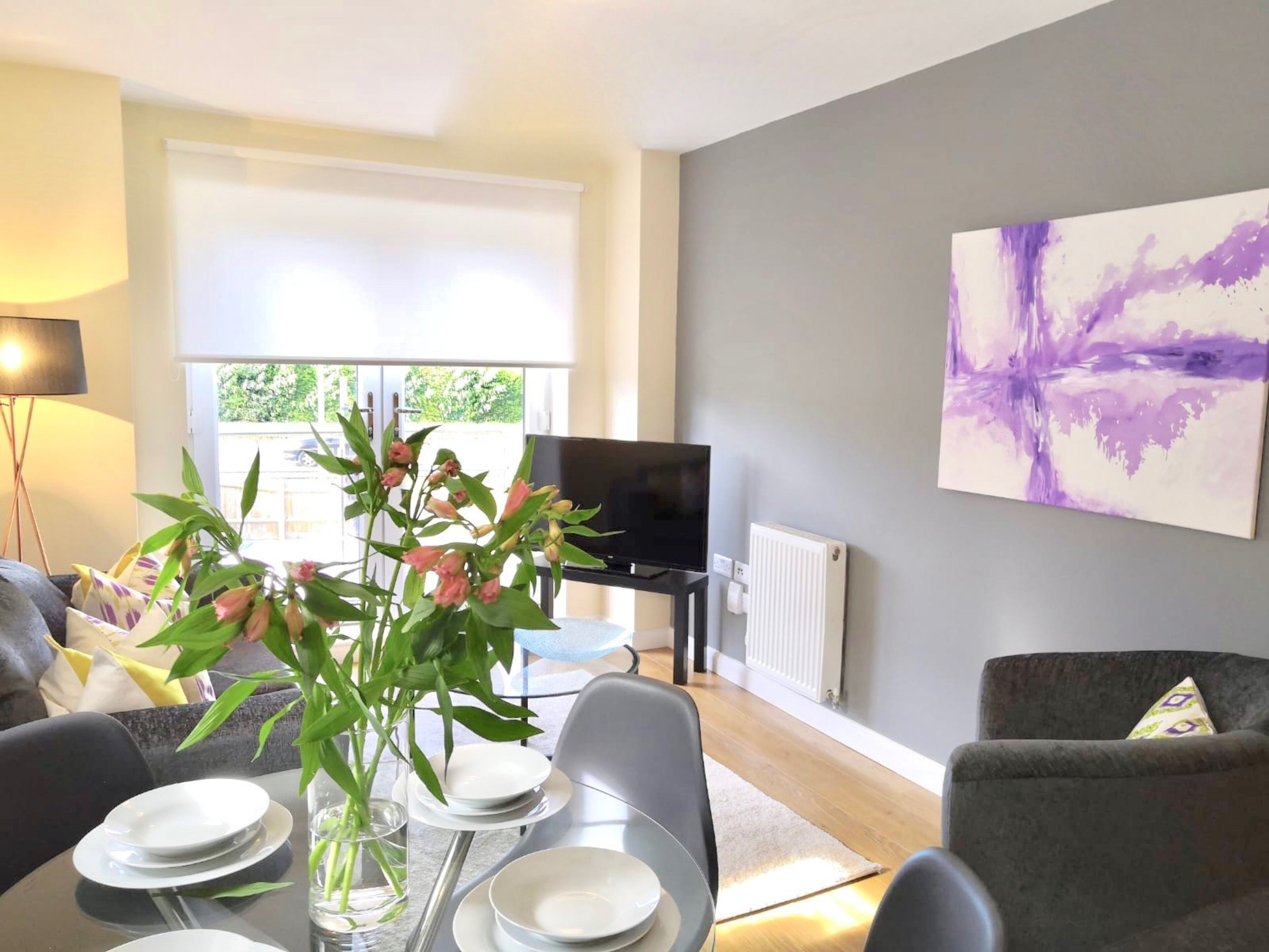 A modern living room with a dining table set for four, a television, and a large abstract painting on the wall.