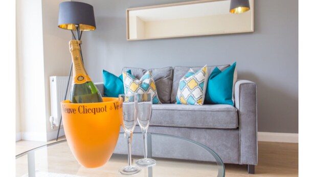 A living room with a grey sofa, colorful cushions, a glass table, a bottle of Veuve Clicquot, and two glasses. - Razia Apartments (Oxford)