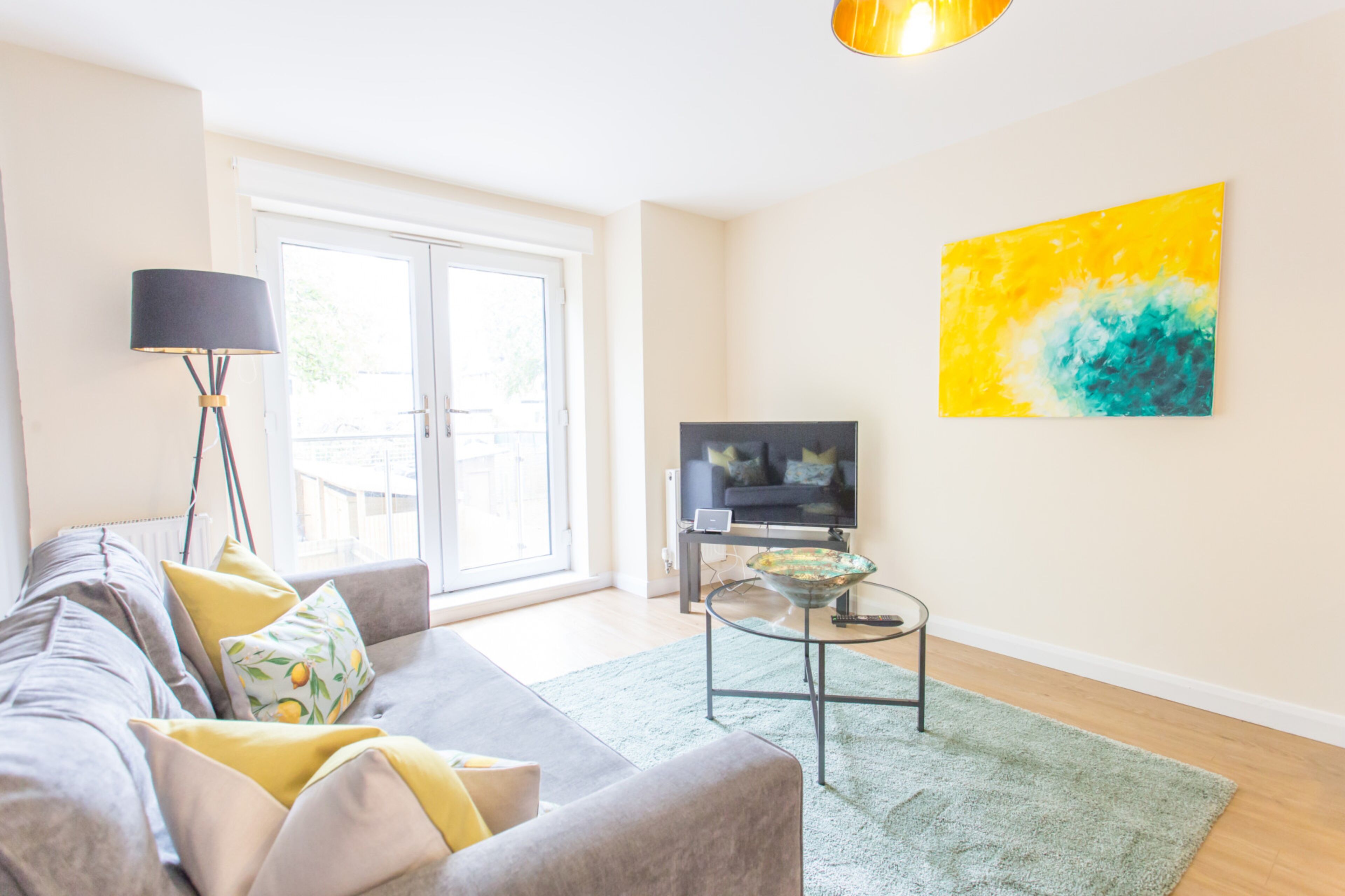 Een moderne woonkamer met een bank, een televisie op een dressoir, een vloerlamp en een groot raam.