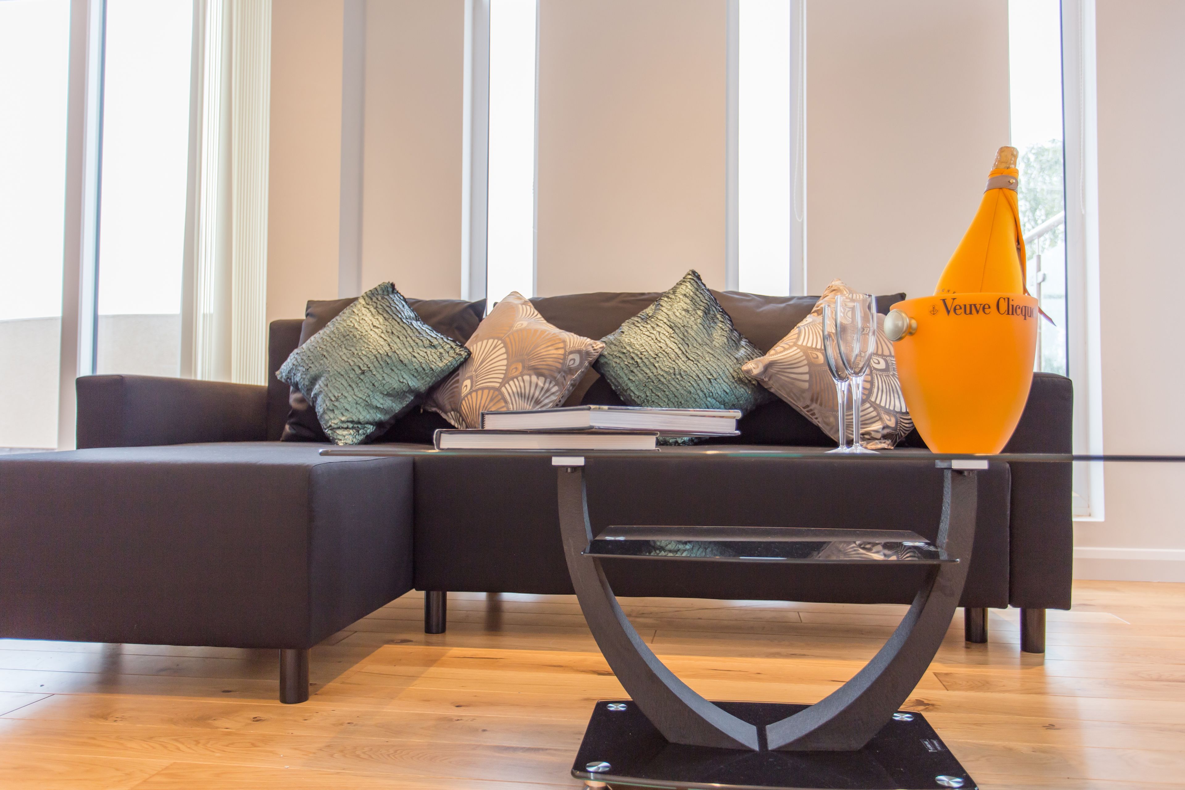 A modern living room with a sectional sofa, decorative pillows, a glass coffee table, and a bottle of Veuve Clicquot champagne.