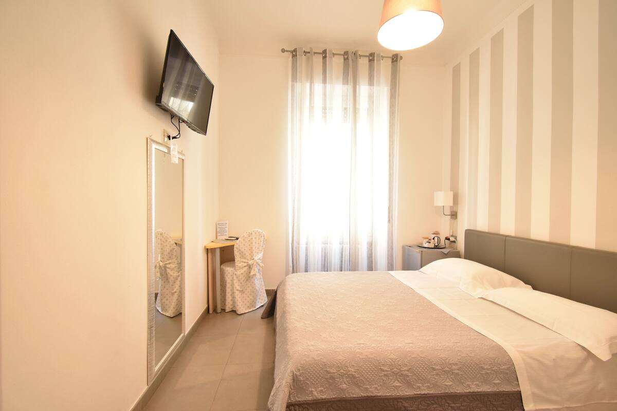 Habitación de hotel con cama, televisión montada en la pared, una silla, mesita de noche y ventana con cortinas.