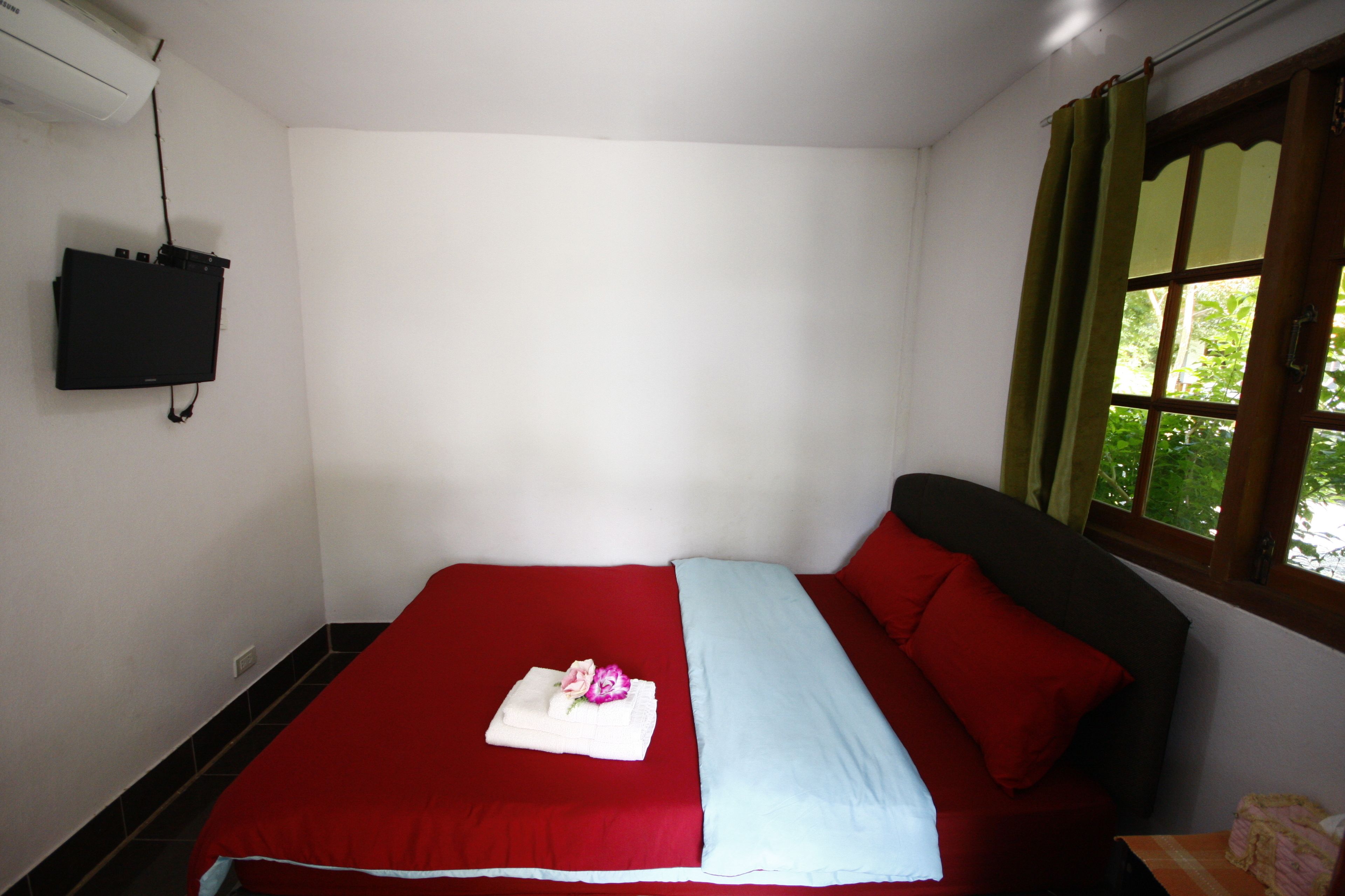 A single bed with a red and blue bedspread, a white pillow, and a folded towel with a flower. There is a window with green curtains and a wall-mounted TV.
