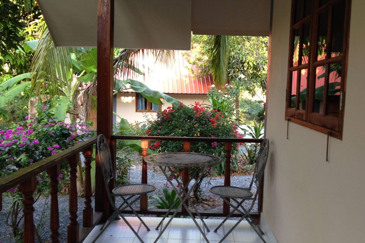 Un patio cubierto con una mesa y dos sillas, rodeado de abundante vegetación y vistas a una casa.