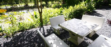 Mesa y bancos de hormigón en un jardín con suelo de grava, rodeado de follaje y árboles.