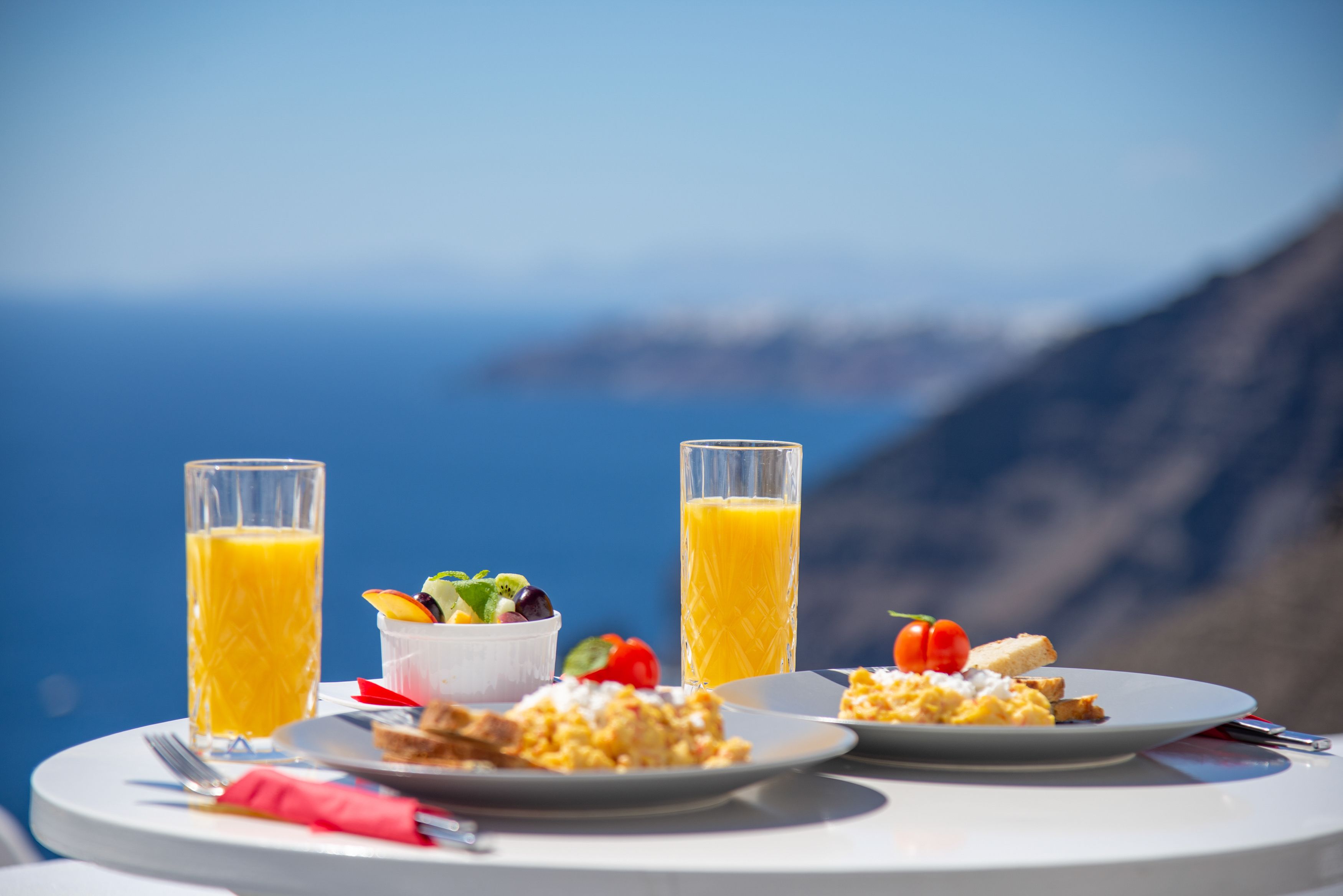 A table with breakfast items including toast, scrambled eggs, and a fruit salad, with two glasses of orange juice, set against a scenic coastal backdrop.