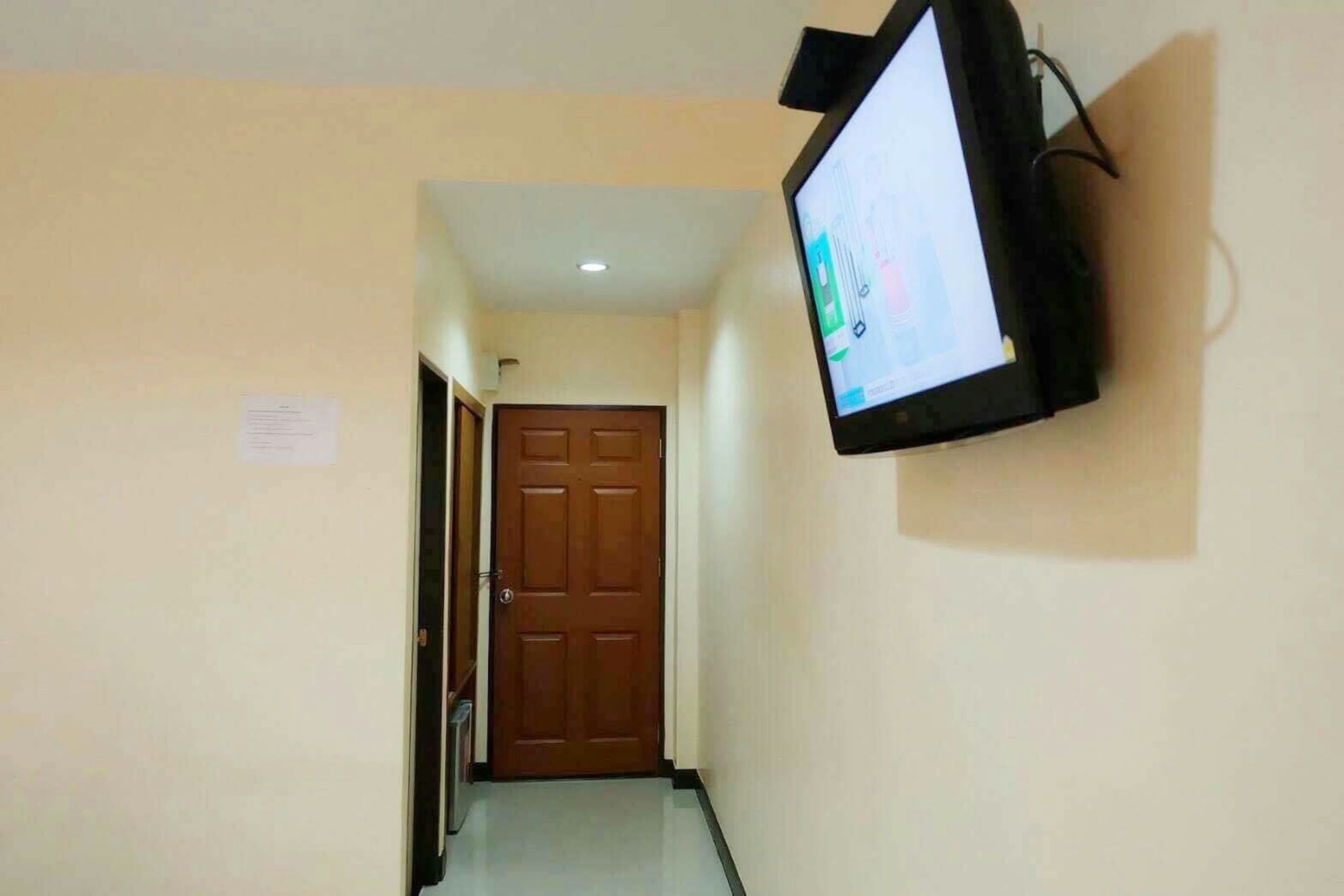 A hallway with a mounted television on the wall, a wooden door at the end, and a light switch on the wall.