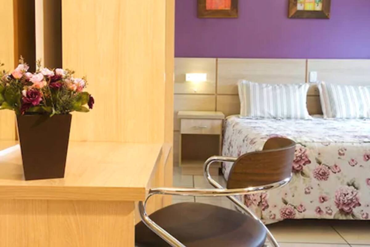 Quarto de hotel com cama, mesa de cabeceira, cadeira, escrivaninha de madeira, planta em vaso e duas molduras na parede.