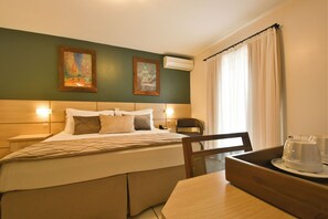 A hotel room with a bed, a desk, a chair, a table, two paintings on the wall, and an air conditioning unit. - Pousada Belluno (Gramado)