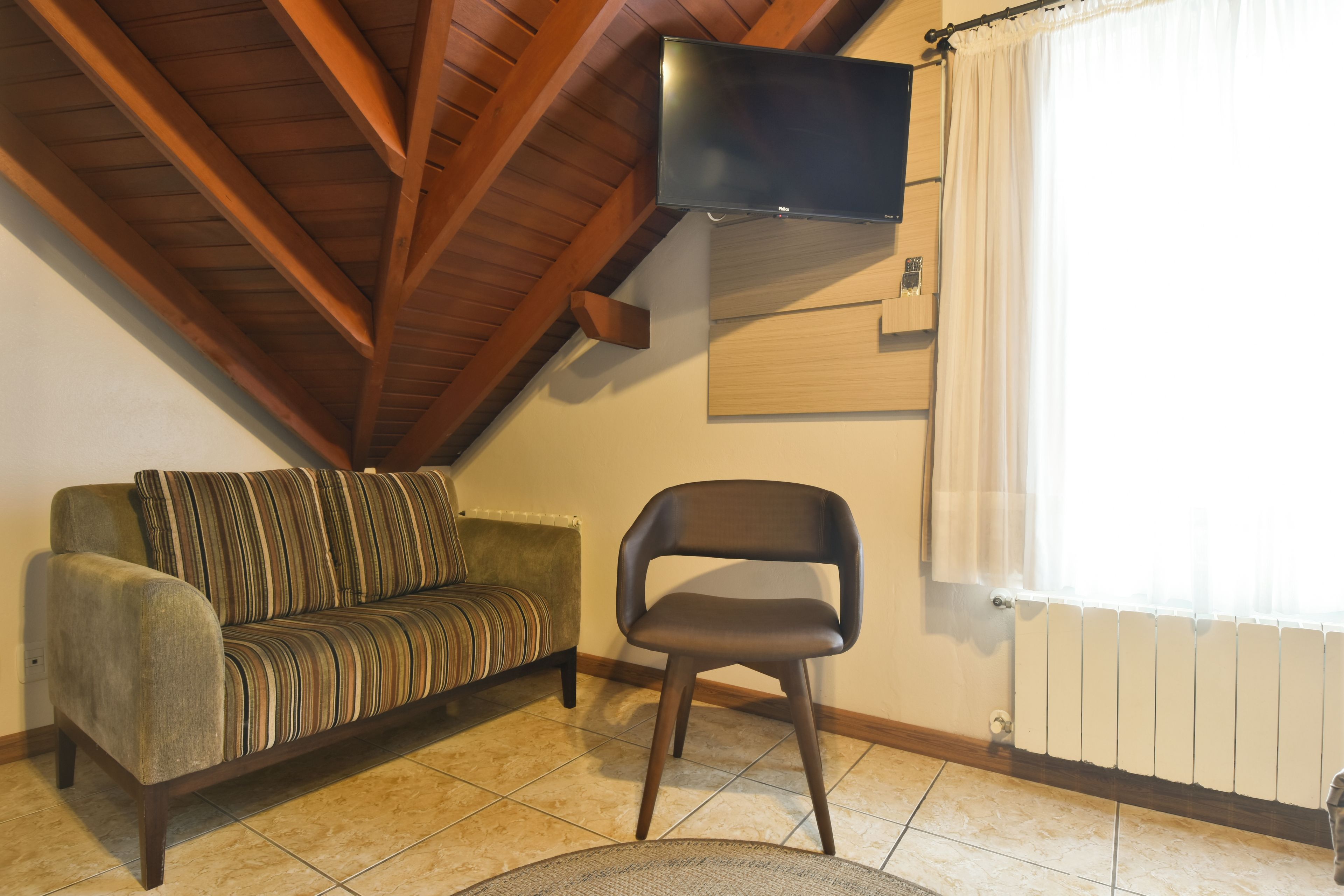 Una sala de estar con techo de madera, un sofá, una silla, un televisor de pantalla plana montado en la pared y una ventana con cortinas.