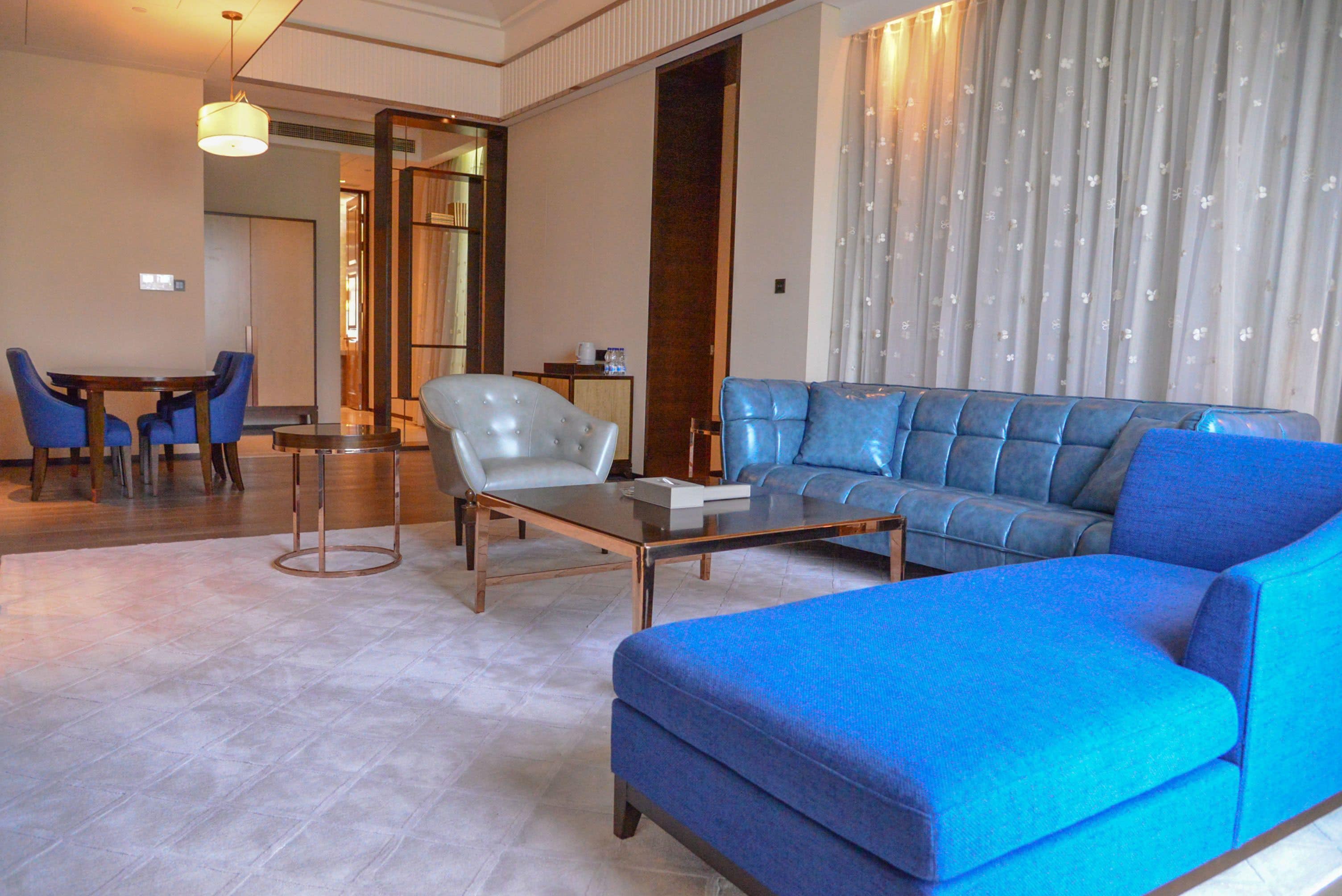 A modern living room with a blue sofa, a leather armchair, and a glass coffee table. There is a dining area with a table and chairs in the background.