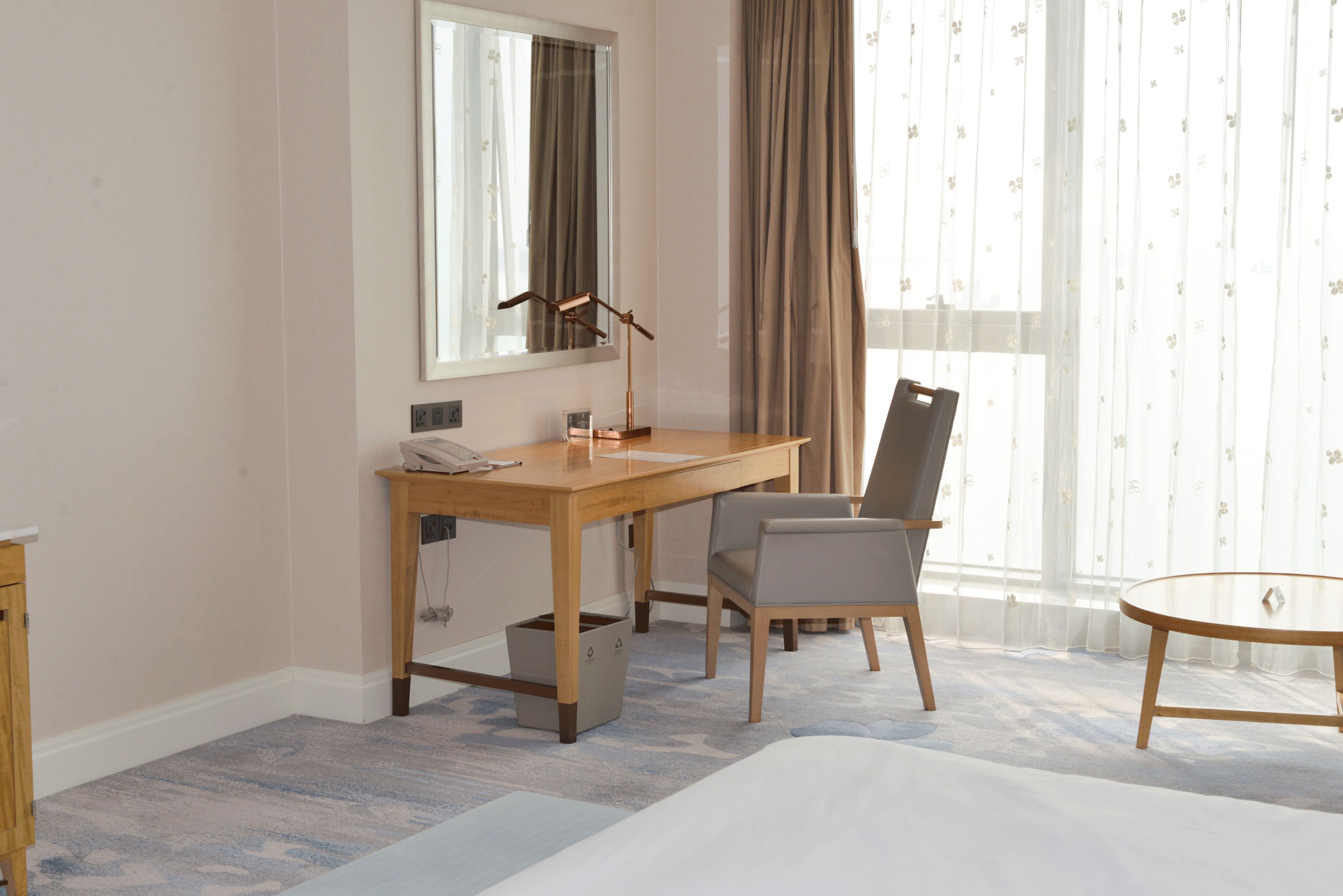 A hotel room with a wooden desk, a chair, a bed, a small table, a mirror, and a window with curtains.