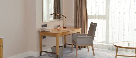 A hotel room with a wooden desk, a chair, a bed, a small table, a mirror, and a window with curtains.