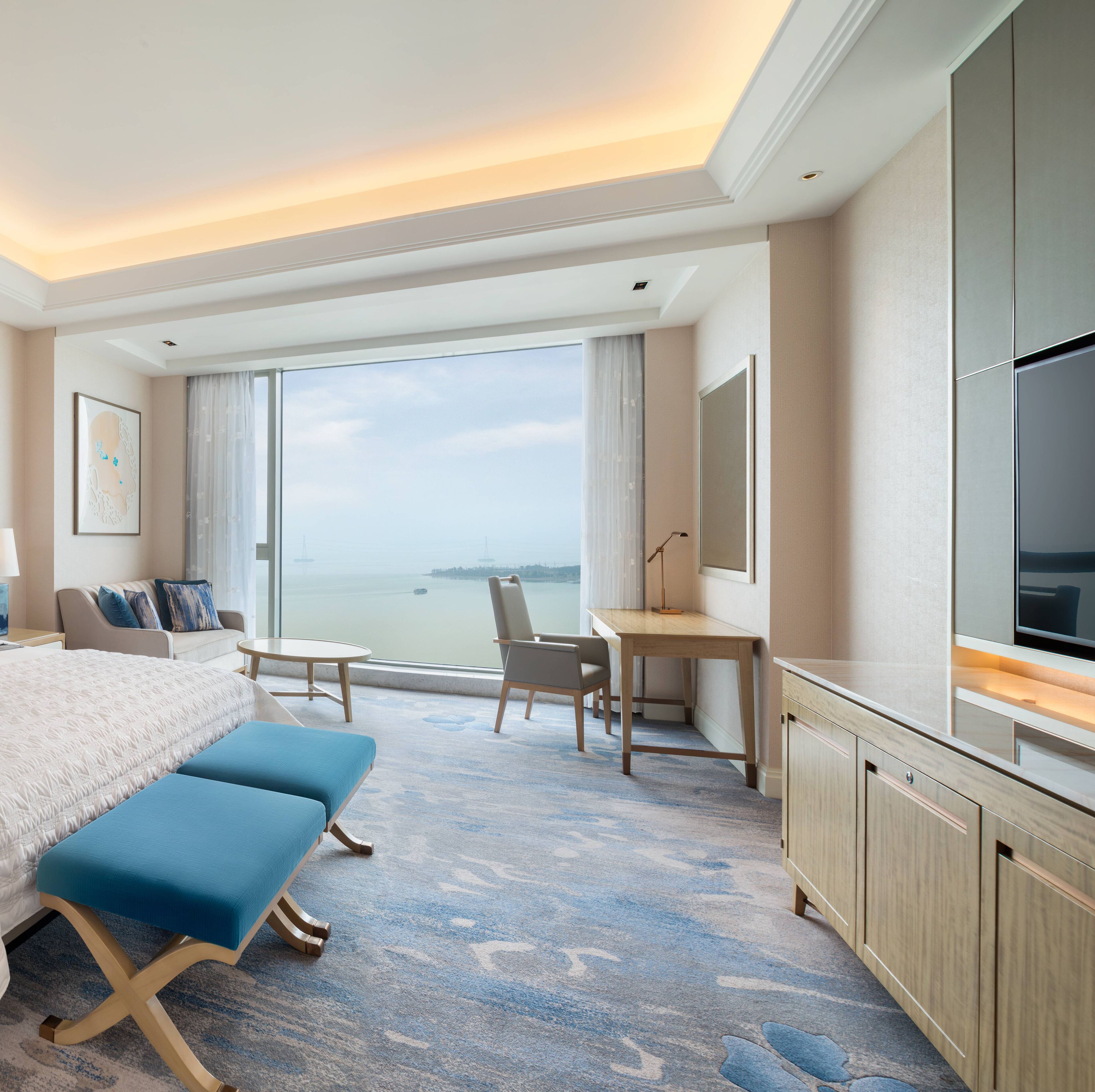 A hotel room with a bed, two blue chairs, a desk, a TV, and a large window offering a view of the sea.