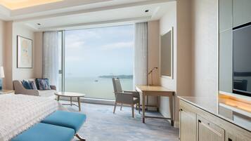 A hotel room with a bed, two blue chairs, a desk, a TV, and a large window offering a view of the sea.