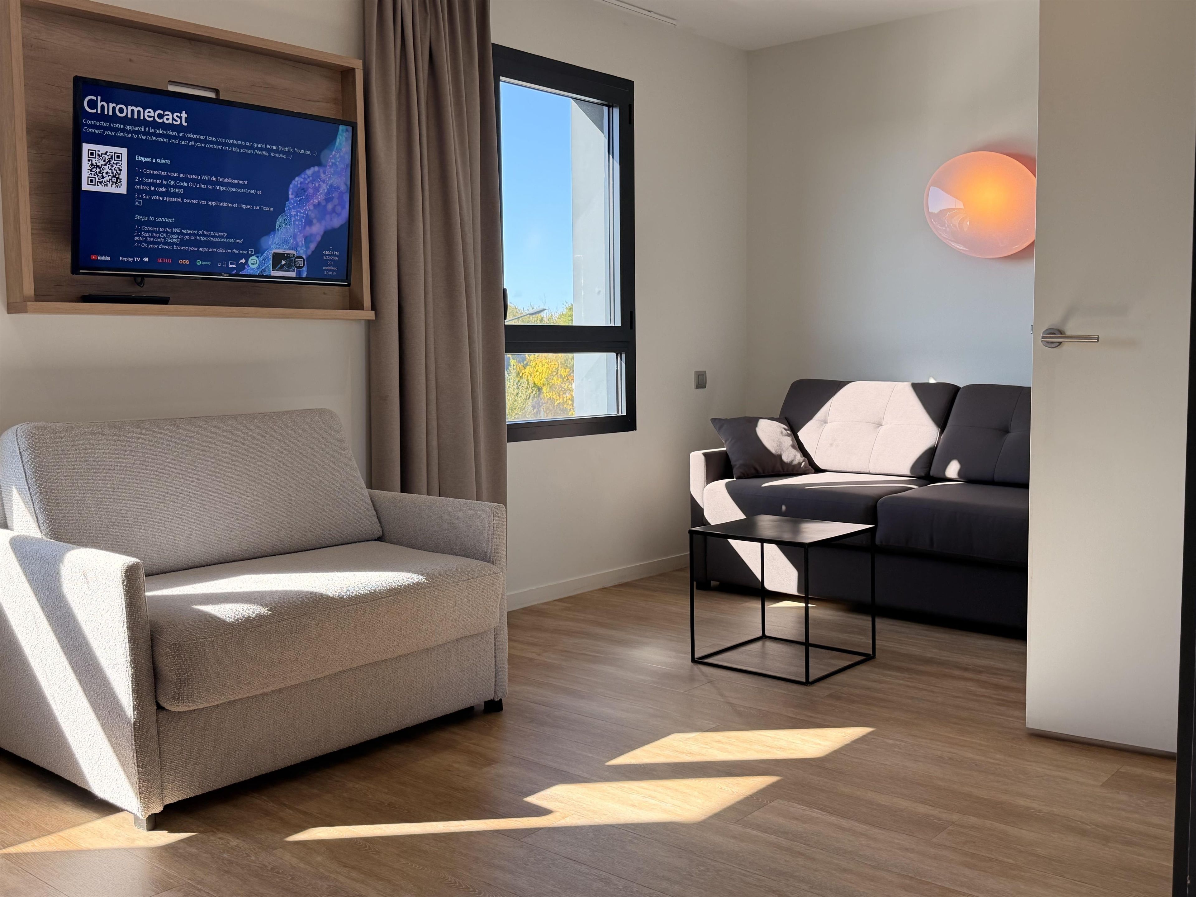 A modern living room with a grey armchair, a black and white sofa, a small black table, and a wall-mounted television displaying a Chromecast setup guide.