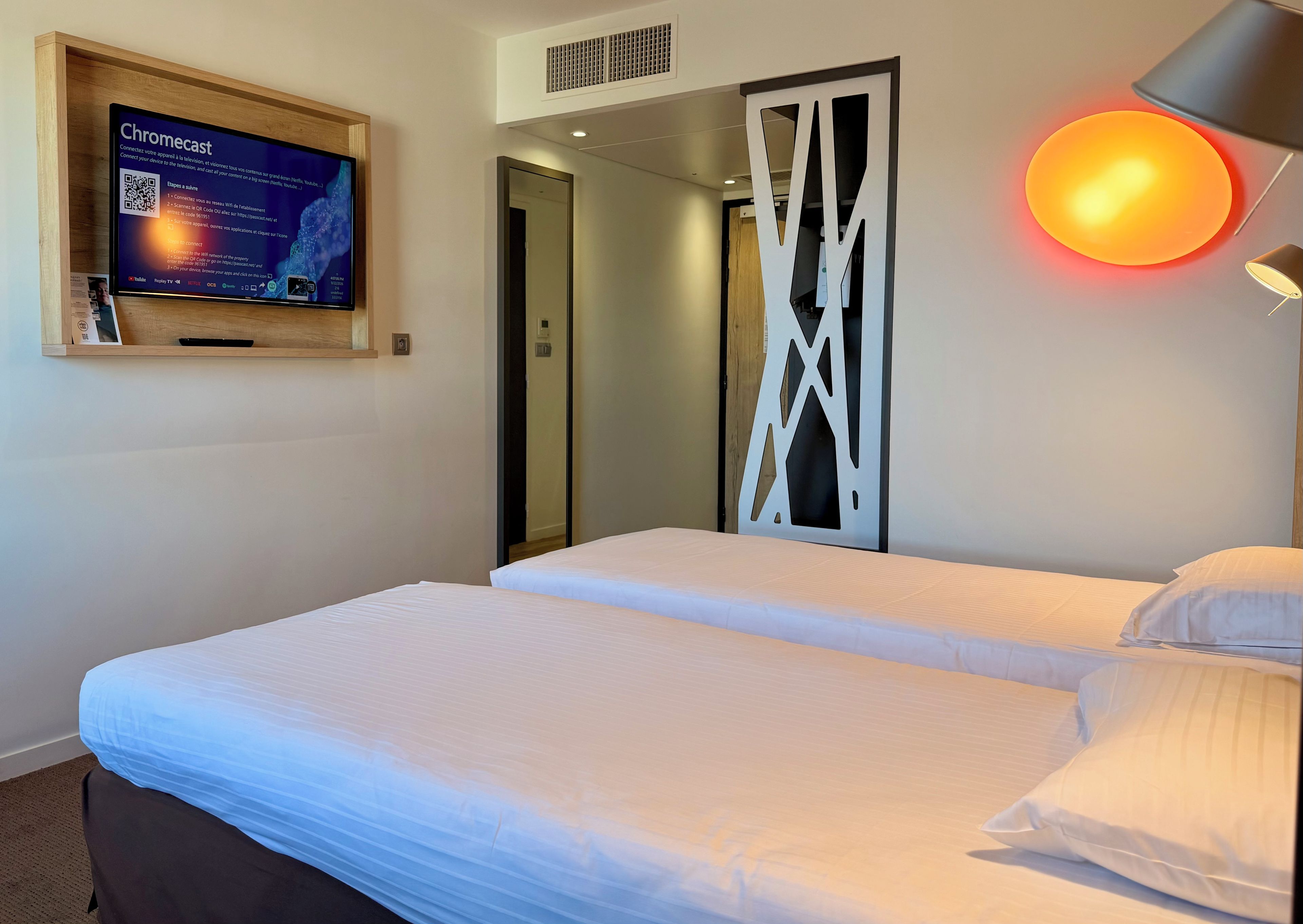 Una habitación de hotel con una cama, un televisor de pared, una lámpara y una puerta.