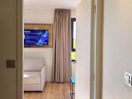 A room with a beige sofa, a television mounted on the wall, a window with curtains, and a door with a handle.