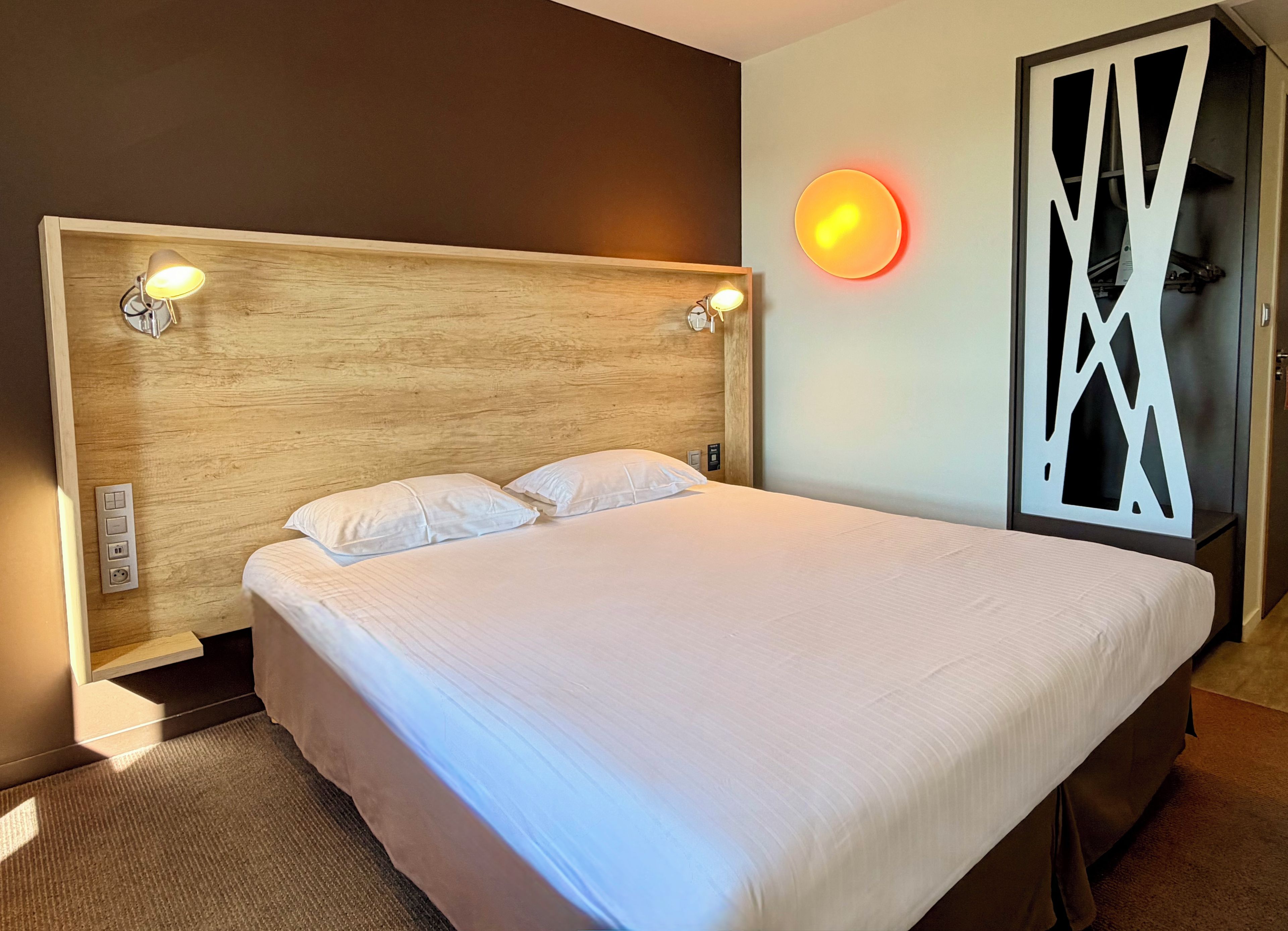 A hotel room with a large bed, two bedside lamps, a wall-mounted light, and a modern decor piece.